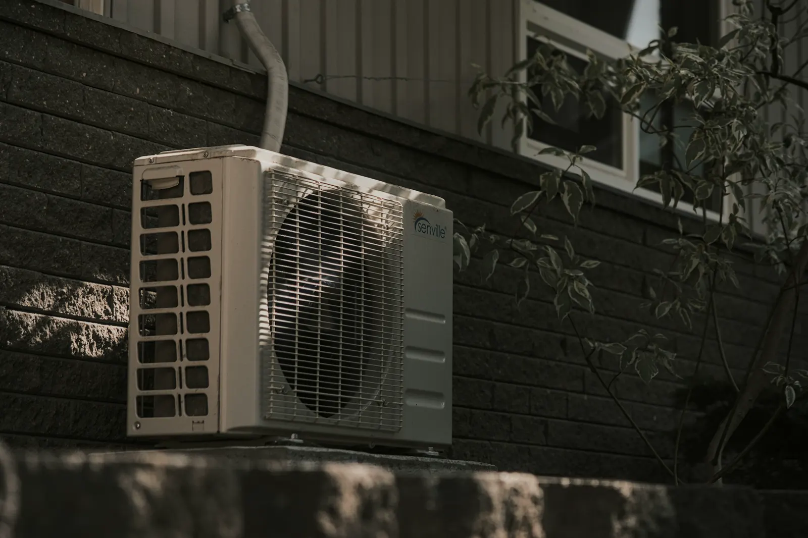 An outdoor heat pump unit on a ledge against a brick wall