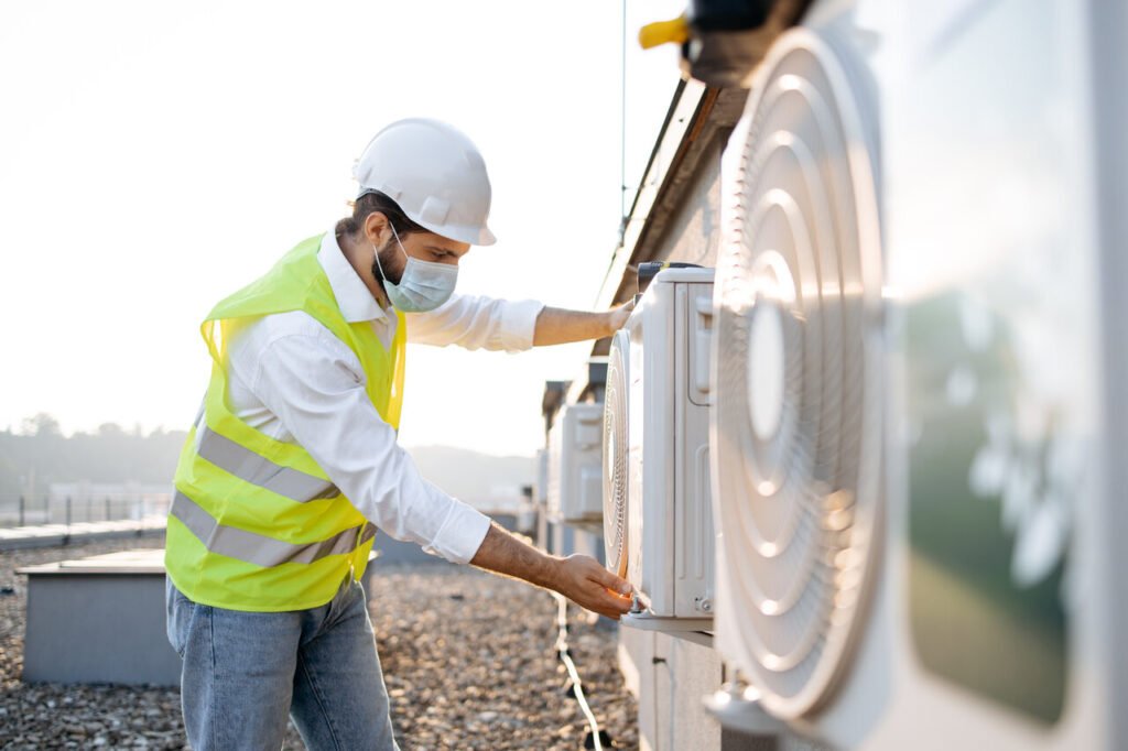 Side view of adult engineer wearing medical mask examining air conditioner while checking work done.